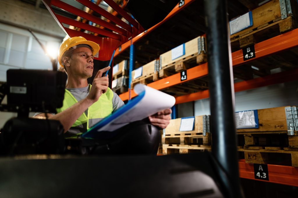 Pensive forklift operator examining stock of packages on shelves in a warehouse.